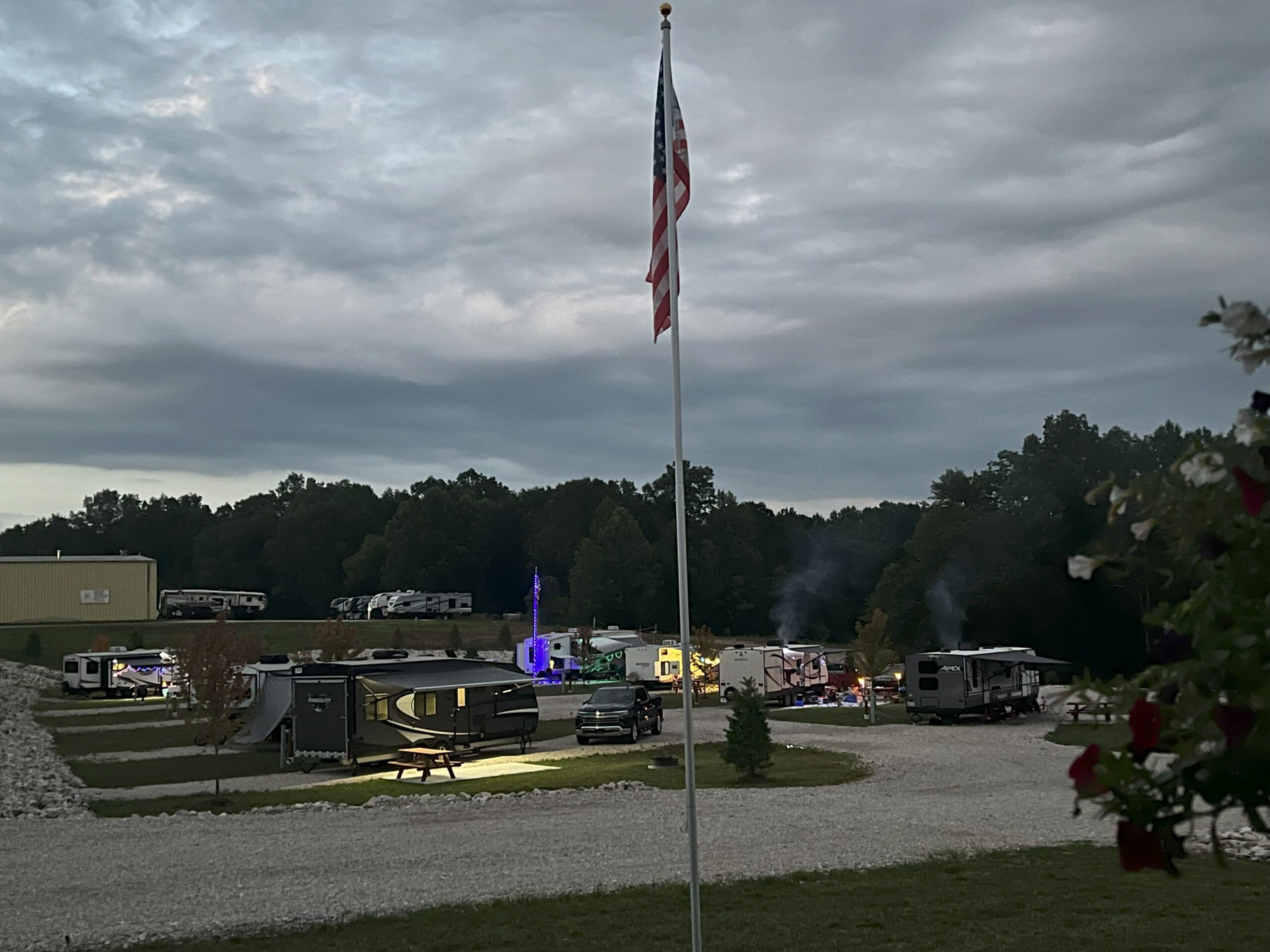 A campground with several RVs parked on gravel lots, a flagpole in the foreground, and trees in the background under a cloudy sky.